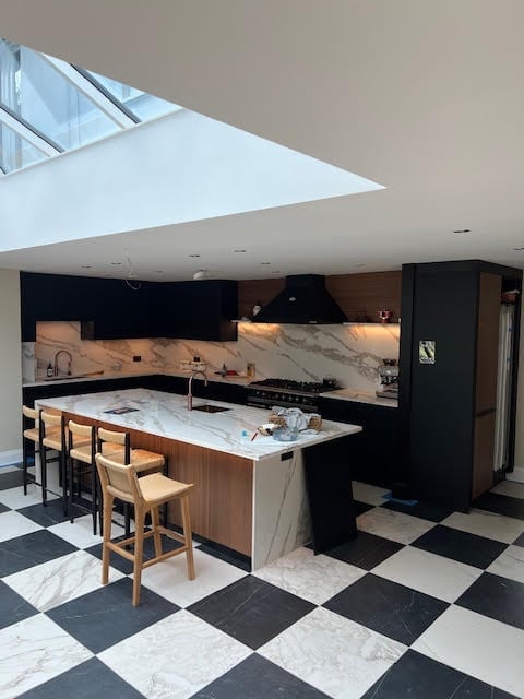 Modern kitchen with black cabinetry, marble countertop island, wooden bar stools, geometric floor, and skylight.