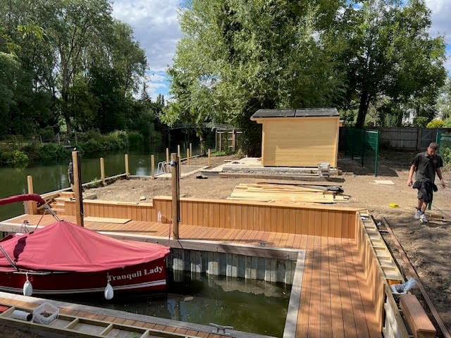 Construction site on a wooden dock beside a canal with a red boat moored nearby and trees in the background
