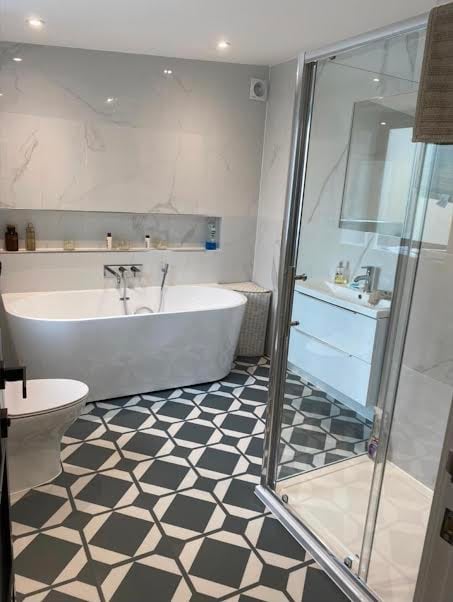 Modern bathroom with white freestanding tub, glass shower enclosure, geometric black and white floor tiles, and marble-effect wall paneling.