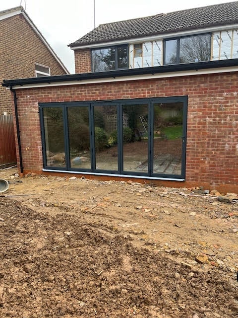 Modern black-framed glass extension with multiple patio doors added to a brick residential house, viewed from bare ground area