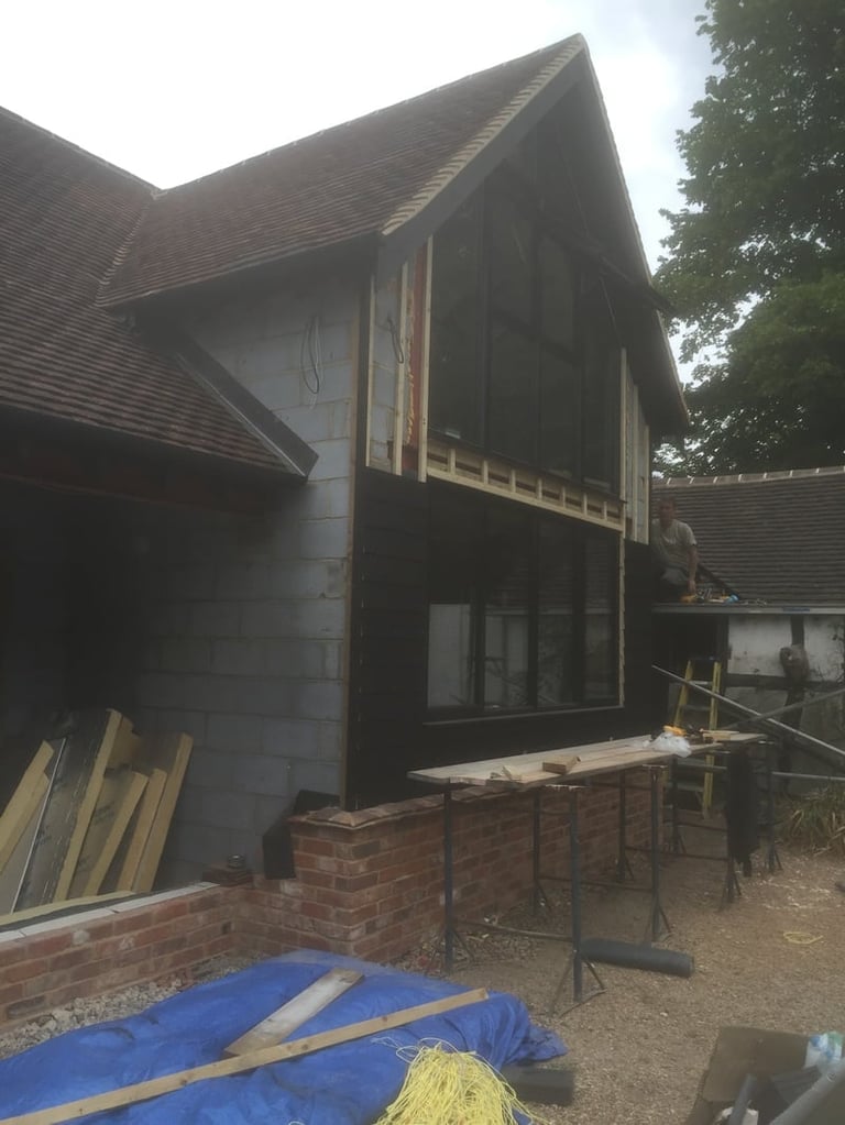 Modern black-clad house extension under construction with brick base, large windows, and building materials scattered around the site