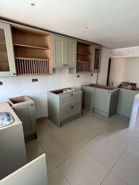 Kitchen under renovation with sage green and white cabinetry, tiled flooring, and open wooden shelving units on the walls