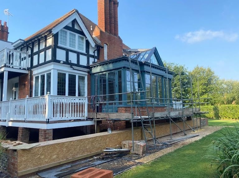 Tudor-style house with brick chimneys and white railings under renovation with scaffolding, surrounded by green lawn and trees