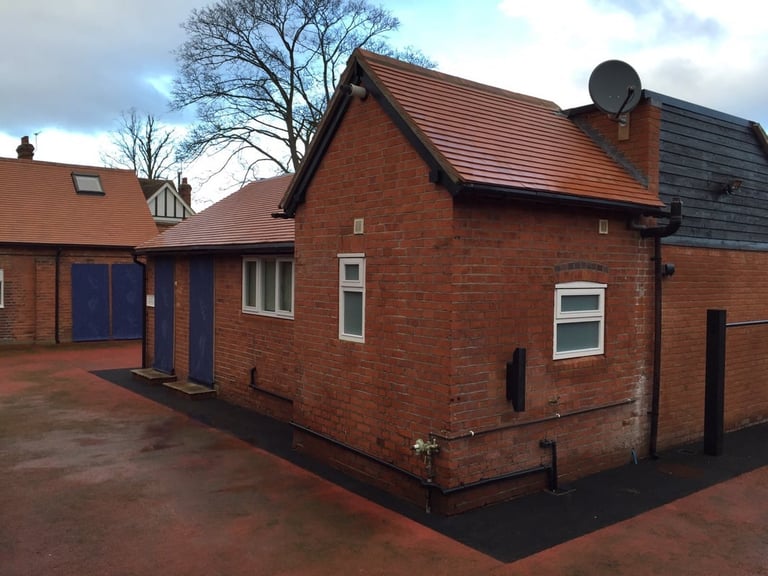 Red brick building with blue door and metal roof, surrounded by paved courtyard and bare tree against cloudy sky