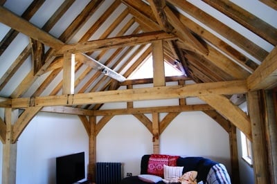 Interior of a rustic wooden barn conversion with exposed timber beams, white walls, and a bed with colorful bedding below