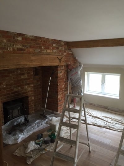 Interior renovation in progress with exposed brick fireplace, wooden beam, ladder, and drop cloths covering the floor and furniture
