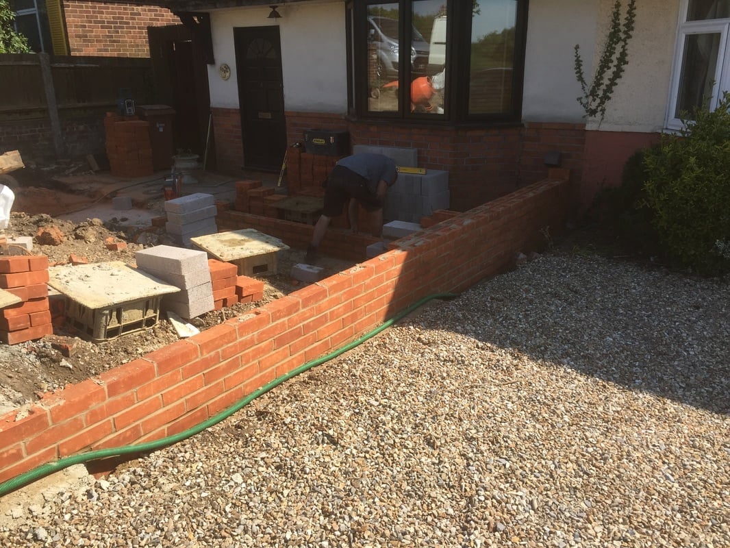 Construction site with red brick wall, gravel ground, and green hose, white house with dark doors in background