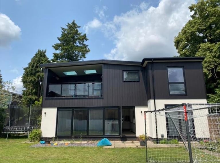 Modern two-story house with black upper section and white lower section, large windows, surrounded by green lawn and trees